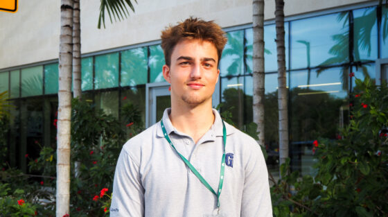 A young man wearing a gray polo shirt and ID badge stands outside in front of plants and a modern building on a sunny day.