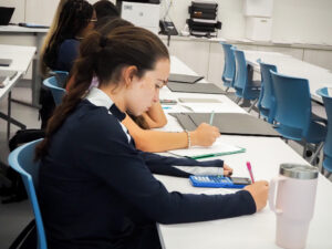 Students sitting at desks in a classroom, writing on paper. One student uses a calculator and has a tumbler on the desk. Blue chairs and office equipment are visible in the background.