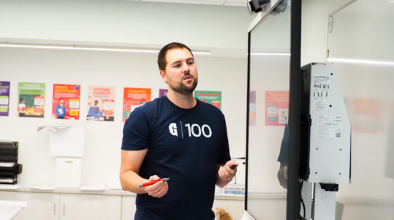 Man wearing a “G 100” shirt stands at a smartboard in a classroom, holding a red marker and a remote, with educational posters visible on the wall behind him.