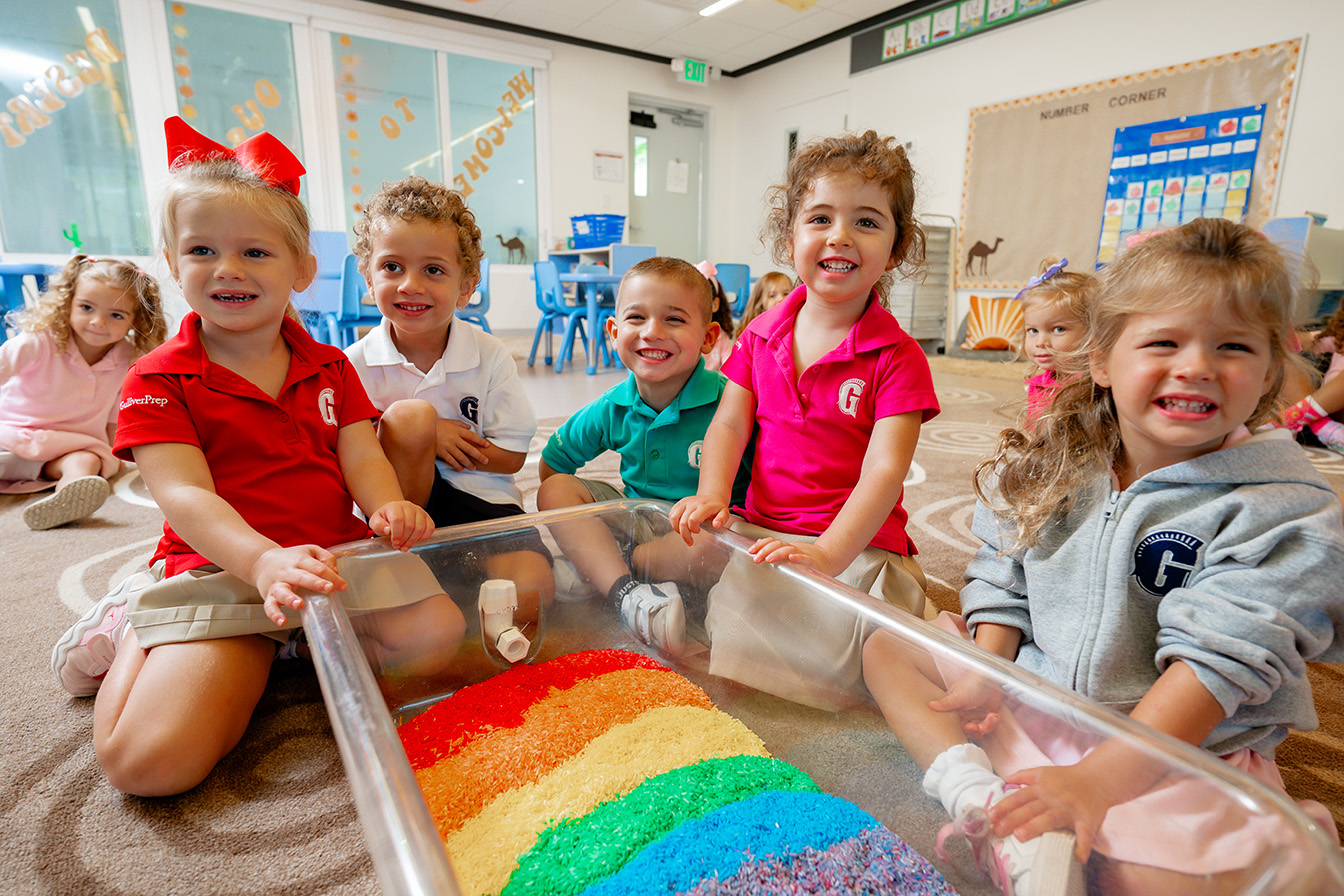 Five young children sit on the classroom floor, smiling around a clear container filled with layers of colorful rice arranged in a rainbow pattern. Other children and classroom decor are visible in the background.
