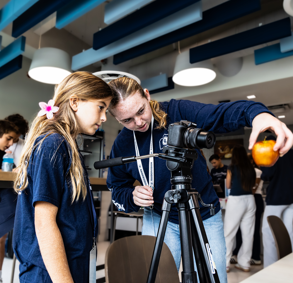 Two girls in navy shirts adjust a camera on a tripod indoors, with one holding an orange object in front of the lens. Other people are seen in the background.