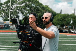 A filmmaker operating a professional movie camera on a football field for an alumni spotlight, with a crew member standing behind him.