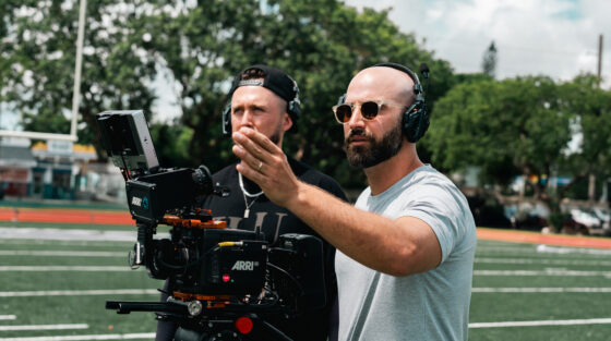 A filmmaker operating a professional movie camera on a football field for an alumni spotlight, with a crew member standing behind him.