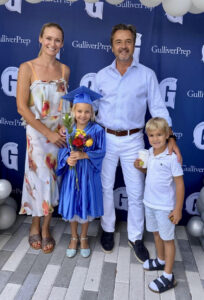 A family poses in front of a Gulliver Prep backdrop. A girl in a blue graduation gown holds flowers, standing with a woman, a man, and a boy.