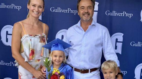 A family poses in front of a Gulliver Prep backdrop. A girl in a blue graduation gown holds flowers, standing with a woman, a man, and a boy.