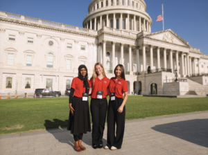 Three young women in red polo shirts with name tags pose for a photo in front of the U.S. Capitol building.