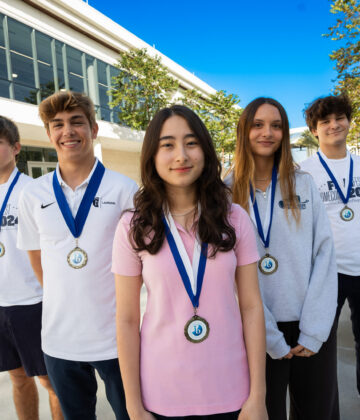 Five students wearing medals stand outside a modern building, posing for a group photo on a sunny day.