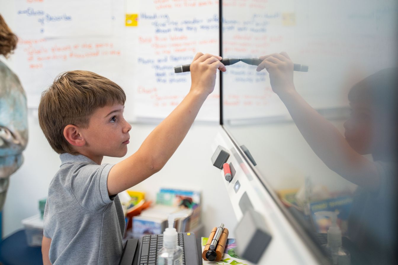 A young boy writes on a whiteboard with a marker in a classroom at a private elementary school, with posters and books visible in the background.