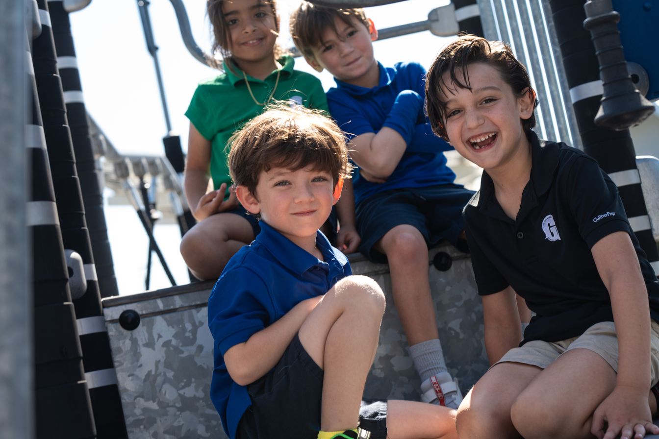 Four children sit and smile together on outdoor playground equipment under bright sunlight.