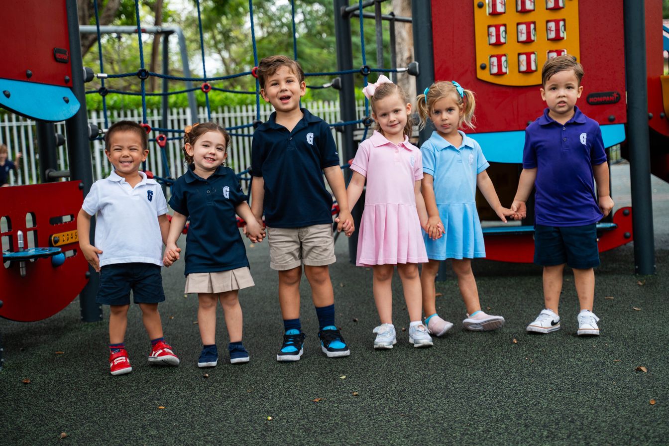 Six young children stand holding hands on a playground, dressed in various colored school uniforms, smiling and facing the camera.