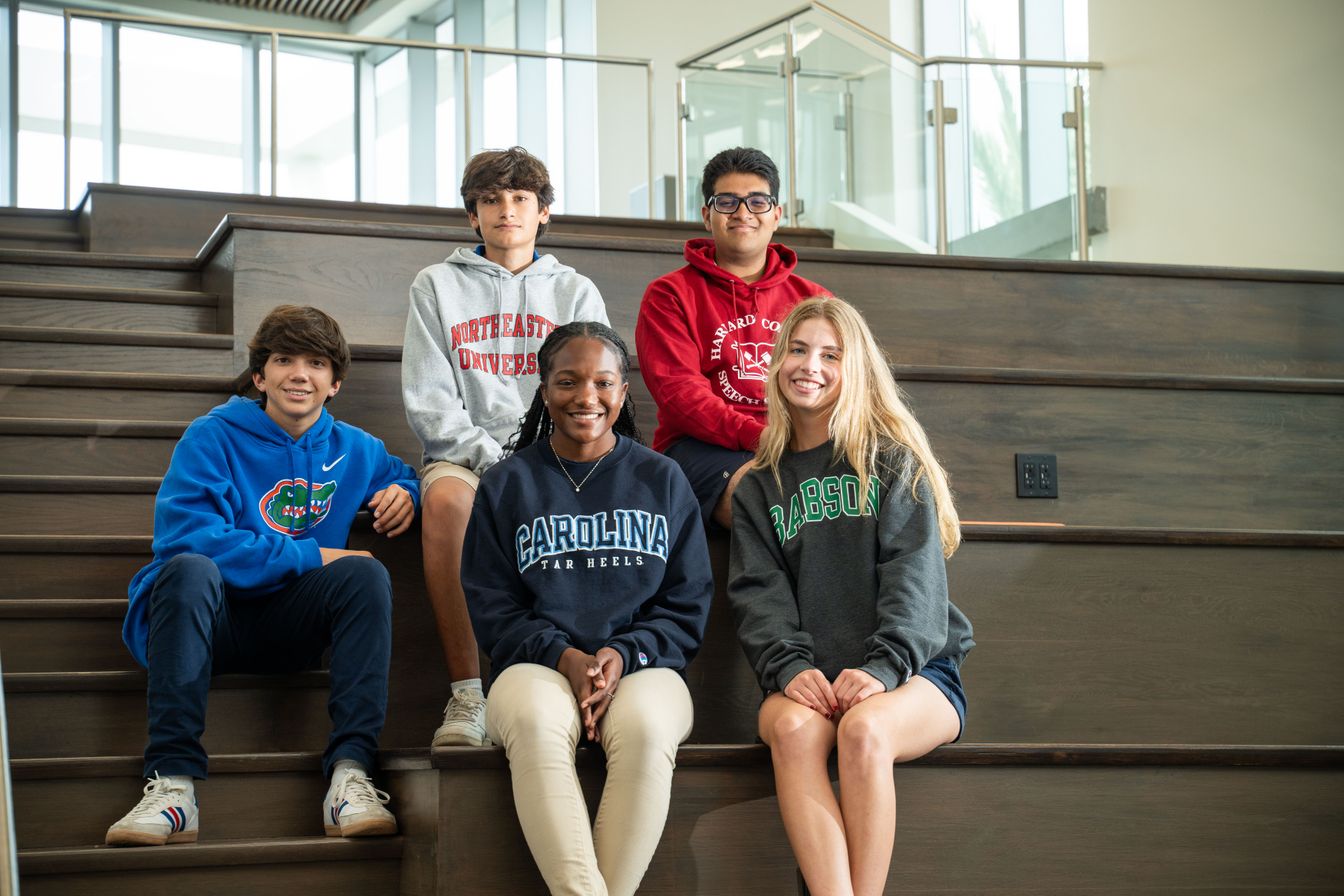 Five students in college sweatshirts sit and stand on wooden stairs in a modern building, smiling at the camera.