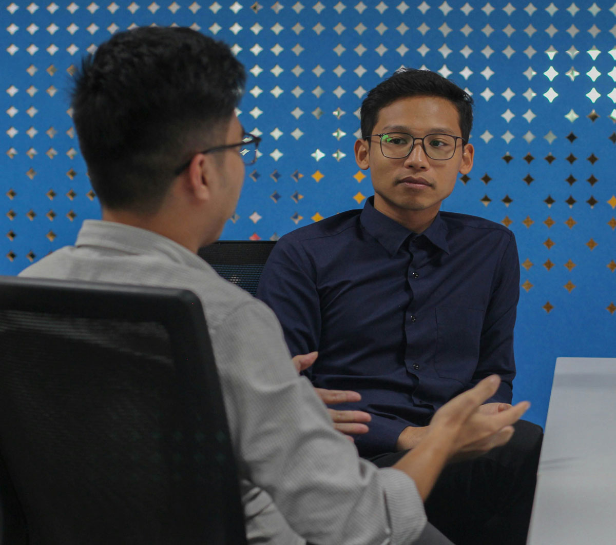 Two men sit at a table having a conversation in an office setting with a blue patterned wall in the background, discussing opportunities and growth within private school careers.