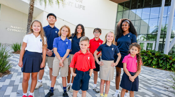 Nine children in school uniforms stand outdoors on a paved area in front of a building with glass windows and greenery.