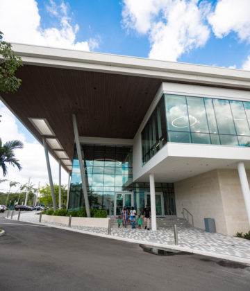 Modern building with large glass windows, overhanging roof, and people standing near the entrance; palm trees and parked cars are visible nearby under a partly cloudy sky.