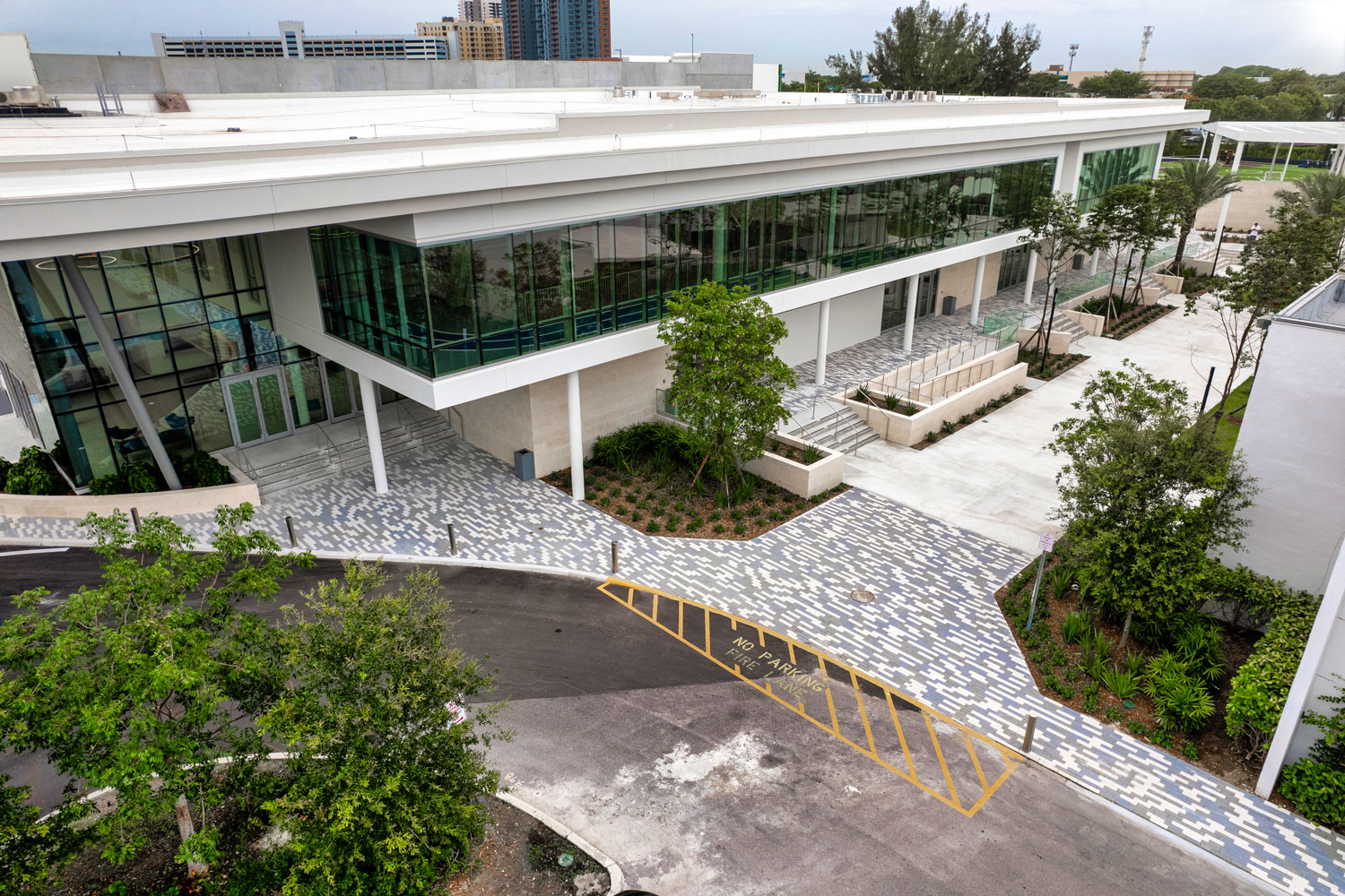 A modern building with glass windows, a covered entrance, patterned walkway, and surrounding trees, viewed from above.