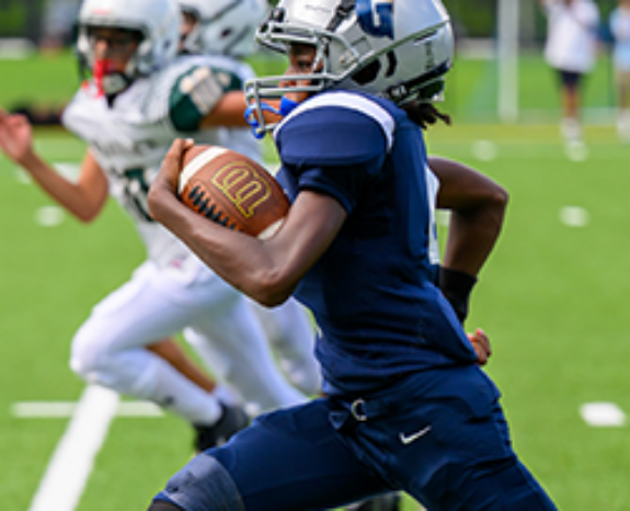 A youth football player in a blue uniform runs with the ball on a green field, while two players in white uniforms chase behind.