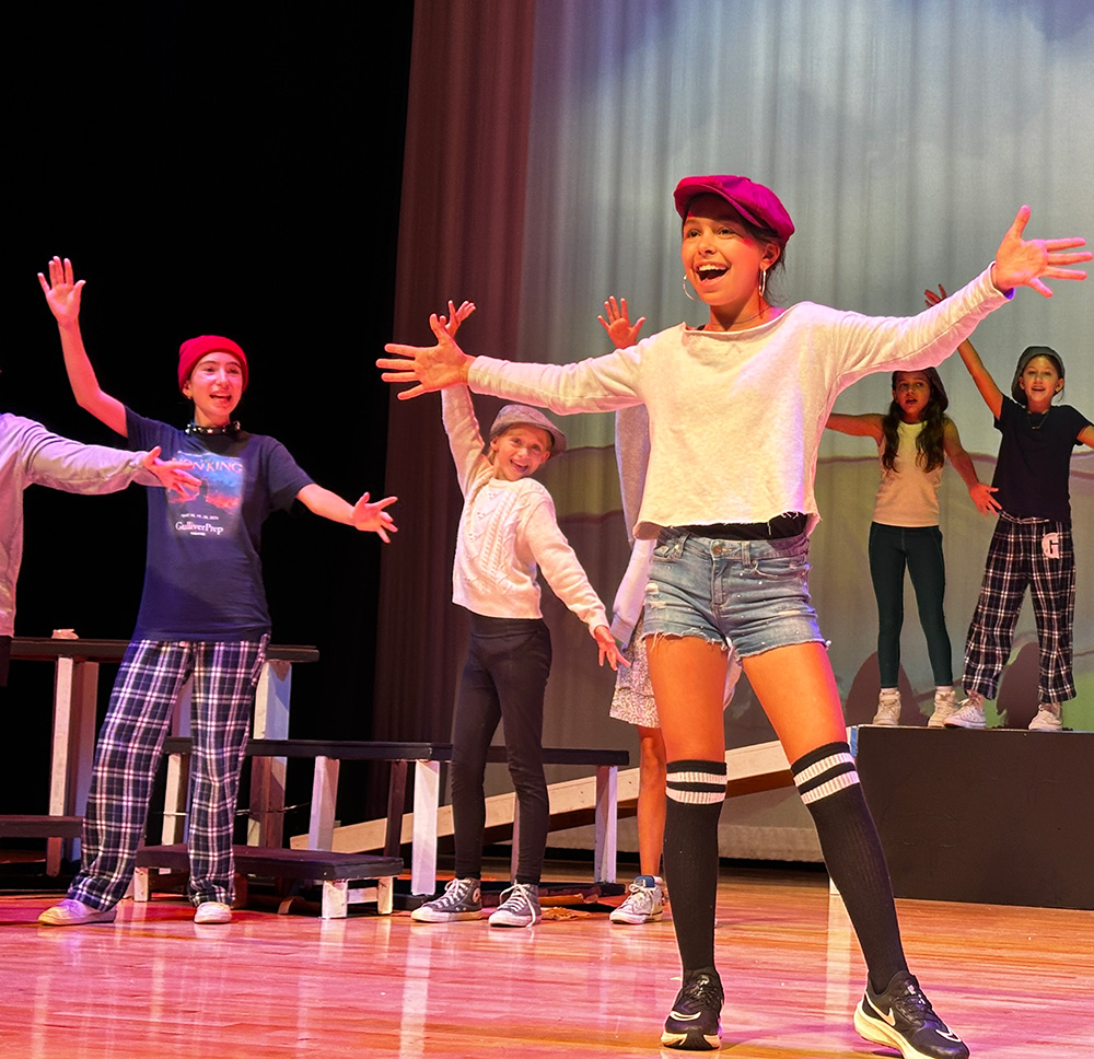 Children perform on stage; one girl in front sings with arms wide, wearing a red cap, white shirt, and shorts. Other kids stand behind her, all with arms raised, acting in a theatrical play.