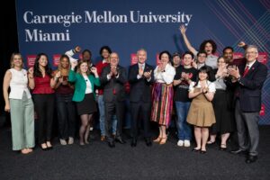 A group of people standing in front of a blue “Carnegie Mellon University Miami” backdrop, with many clapping and smiling.