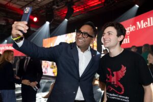 A man in a blazer and a young man in a Carnegie Mellon University t-shirt taking a selfie together at an event.