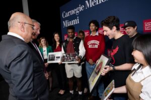 A group of students presenting framed artwork to two men in suits at a Carnegie Mellon Miami event.