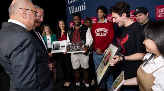 A group of students presenting framed artwork to two men in suits at a Carnegie Mellon Miami event.