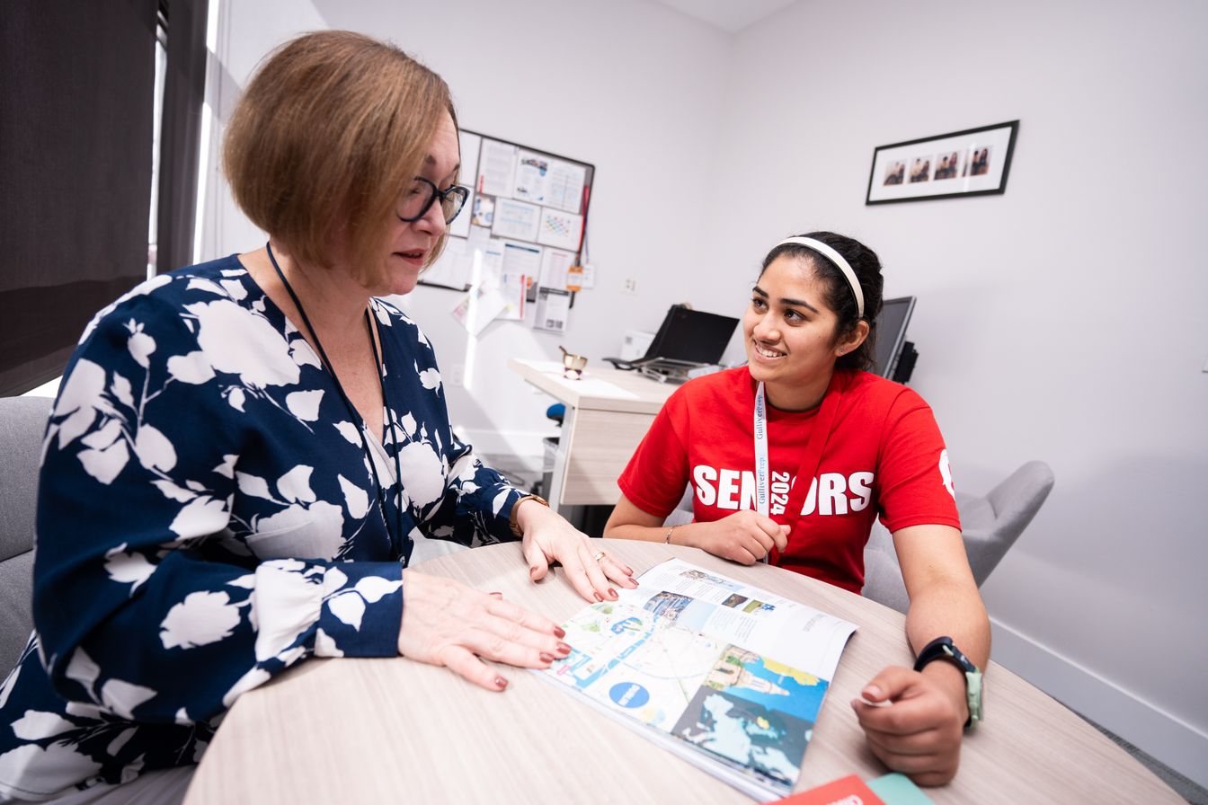 Two women sit at a table in an office, looking at papers and maps together. One wears a floral blouse, the other a red “Seniors 2024” shirt.