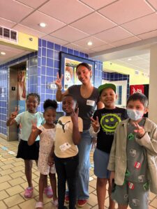 A young woman stands behind five smiling children in a lobby, all making peace signs with their hands.