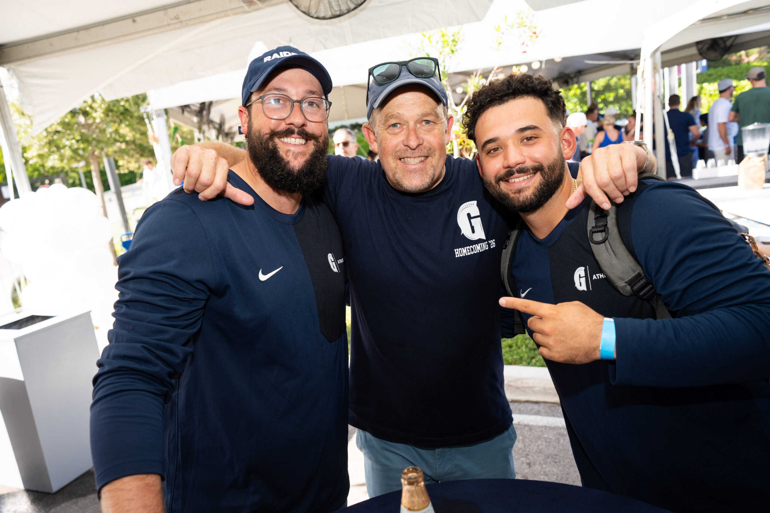 Three men in matching navy blue long-sleeve shirts stand together smiling at a private school careers event.