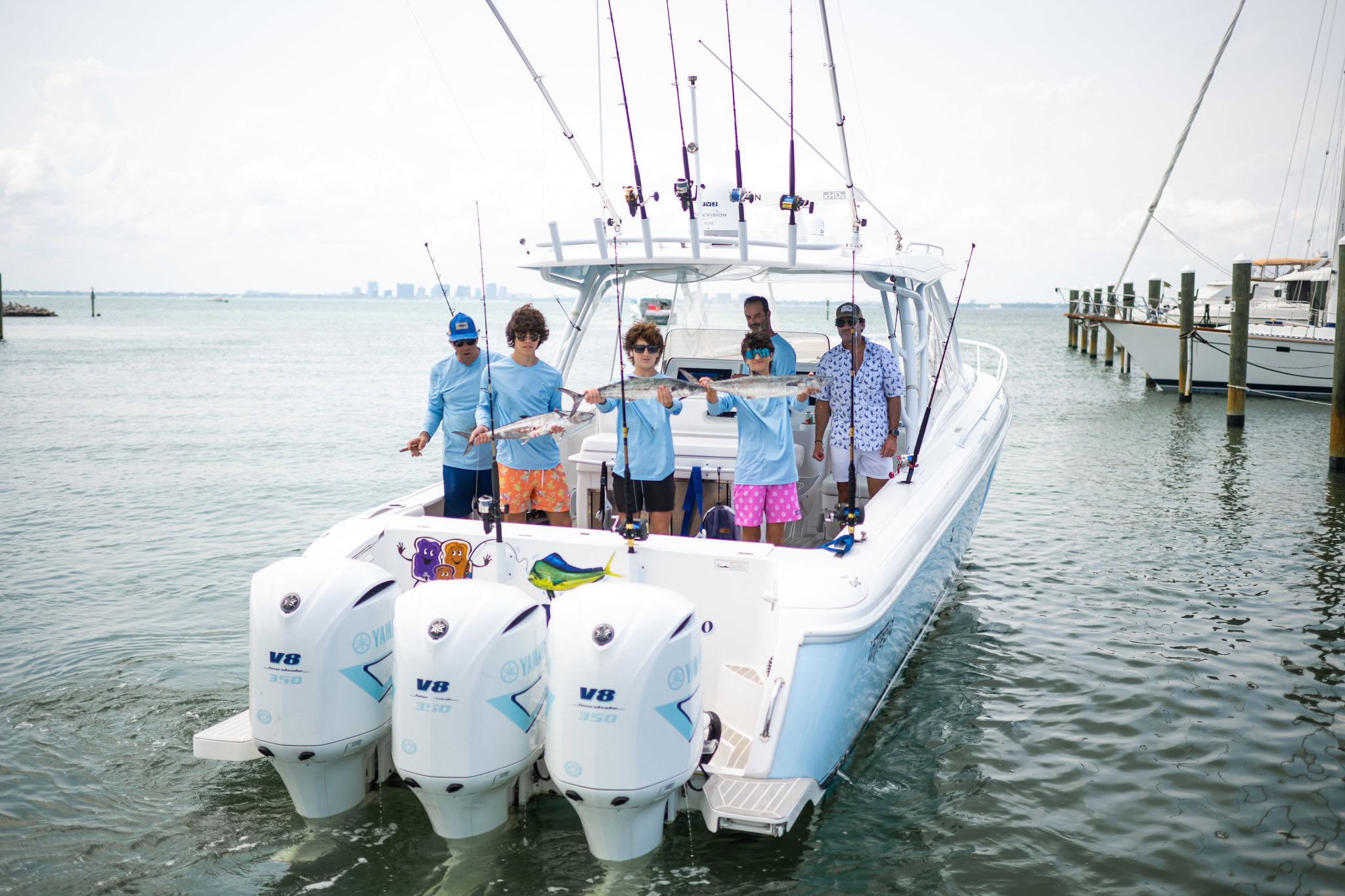 Six people stand on a docked fishing boat; two children at the center display a caught fish. The boat is equipped with three large outboard motors and multiple fishing rods.