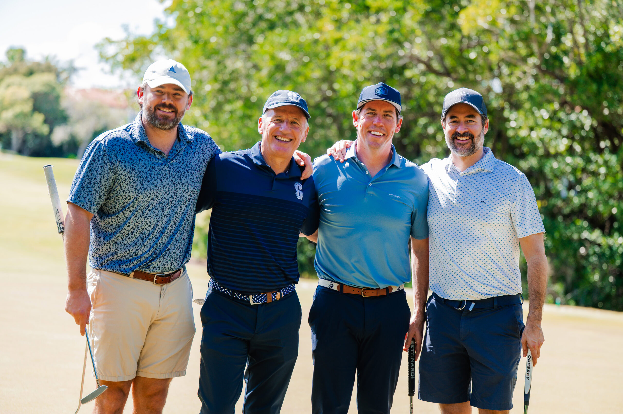 Four men stand together on a sunny golf course, smiling at the camera, each holding a golf club. Trees and greenery are visible in the background.