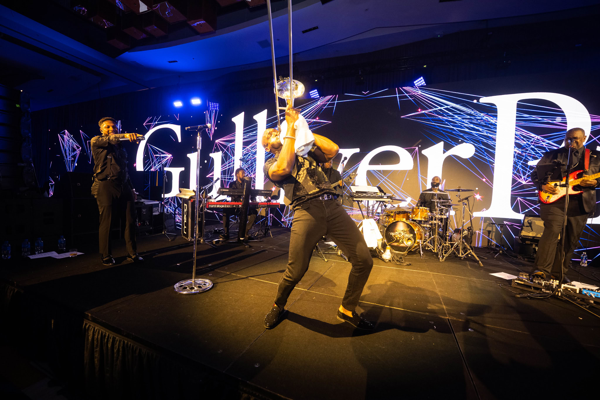 A band performs energetically on stage with a trumpet player in front, vivid stage lights, and “Gulliver” partially visible on the screen behind them.