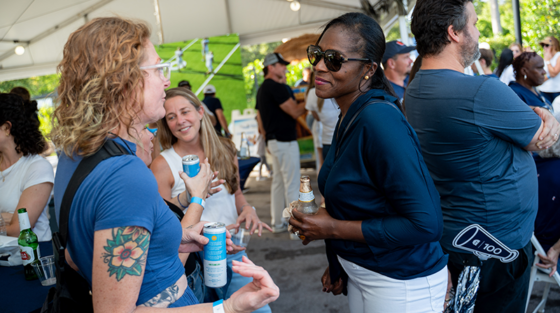 Two women in casual clothing talk while holding canned drinks at a Gulliver Centennial 100 Years outdoor event under a white tent.