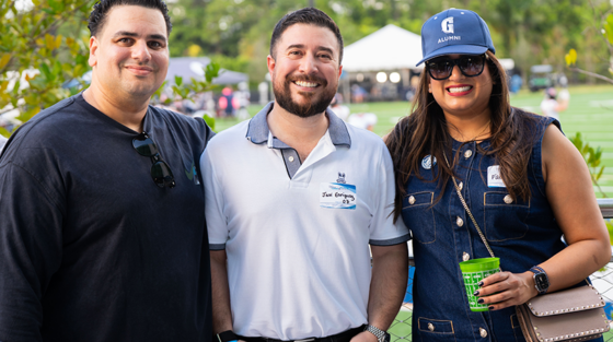 Three people in casual clothing with name tags pose outdoors at a Gulliver Centennial alumni event.