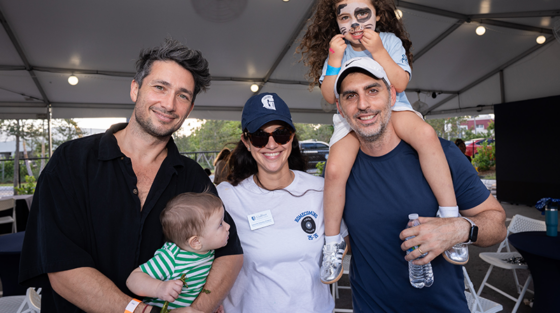 Four adults and two children pose together at an outdoor event under a tent during the Gulliver Centennial 100 Years celebration; one child, with face paint, sits on an adult’s shoulders—a moment perfect for the Alumni Newsletter.