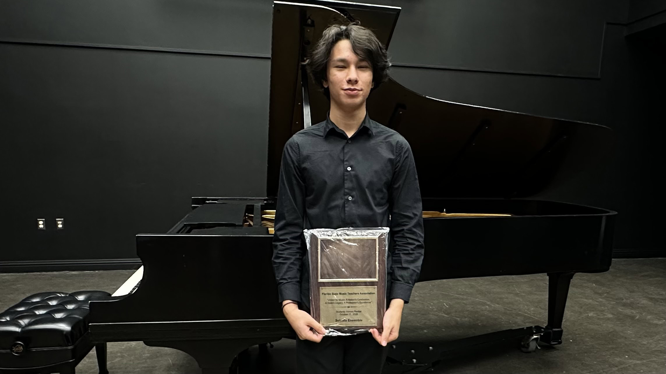 A person in a black button-down shirt stands in front of a grand piano holding a framed plaque.