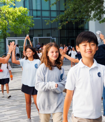 Students in uniforms walk outside a school building, smiling, while adults on either side give high fives.