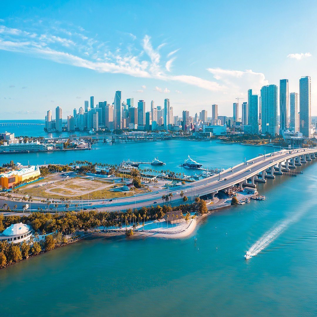 Aerial view of the Miami skyline across Biscayne Bay, a coastal area where many professionals pursue private school careers, featuring a bridge and a boat leaving a wake.