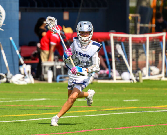 Lacrosse player in white uniform and helmet runs on a field with a lacrosse stick, while other players and sports equipment are visible in the background.