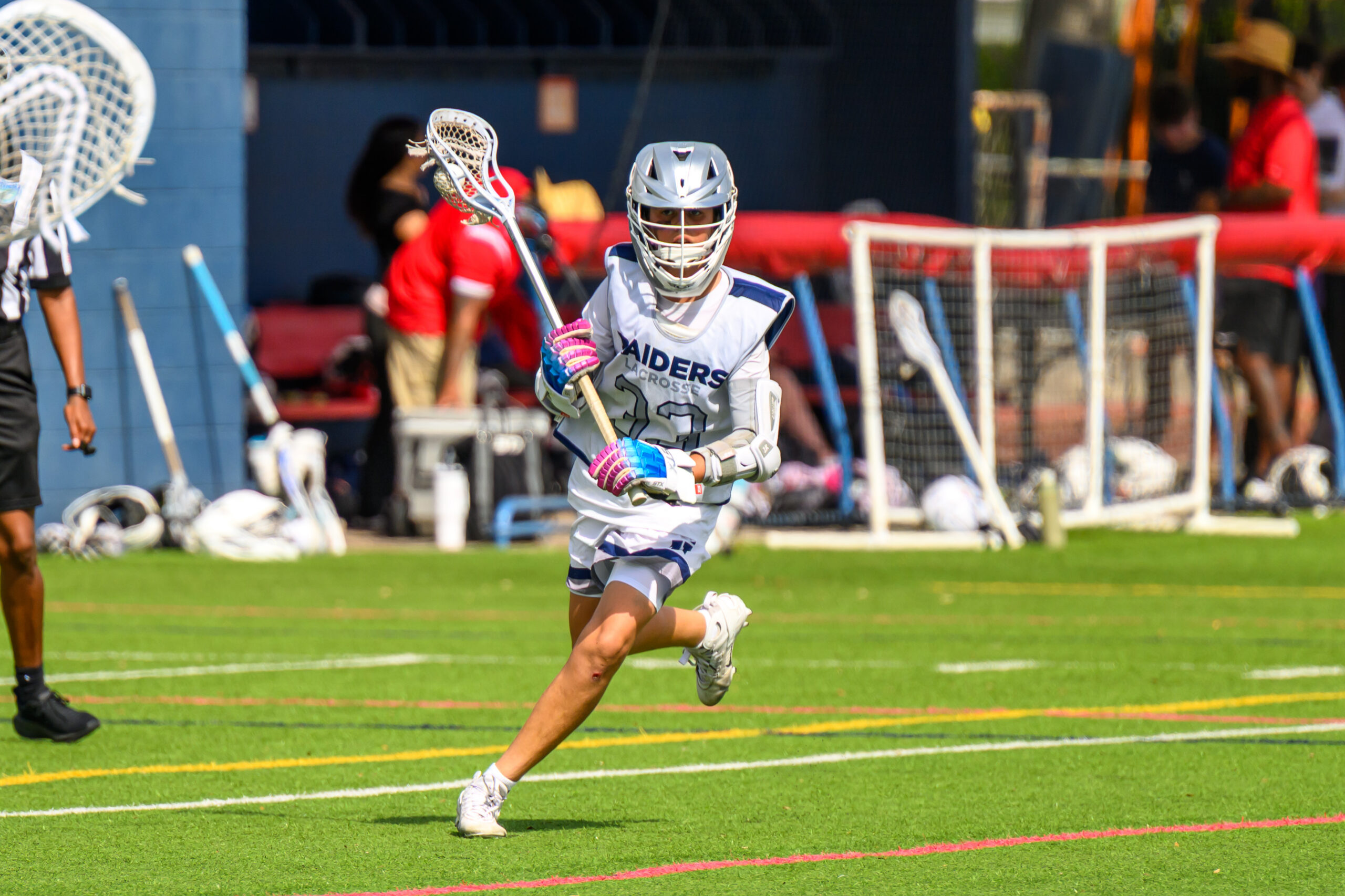 Lacrosse player in white uniform and helmet runs on a field with a lacrosse stick, while other players and sports equipment are visible in the background.