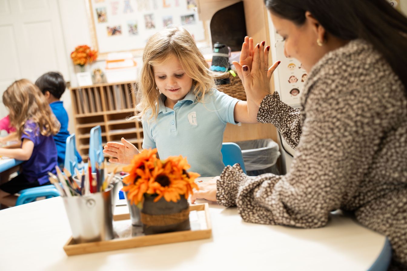 A teacher and a young girl sit at a table in a primary prep classroom, smiling and giving each other a high-five. Other children are working in the background. A vase of flowers is on the table.