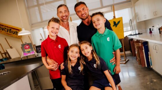 Two adults and four children pose together in a bright art classroom. The children wear school uniforms and smile at the camera. Art supplies and paintings are visible in the background.