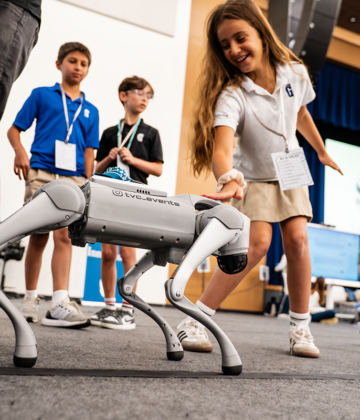 Children interact with a four-legged robot dog on a stage during an educational event. Other students stand and watch in the background.