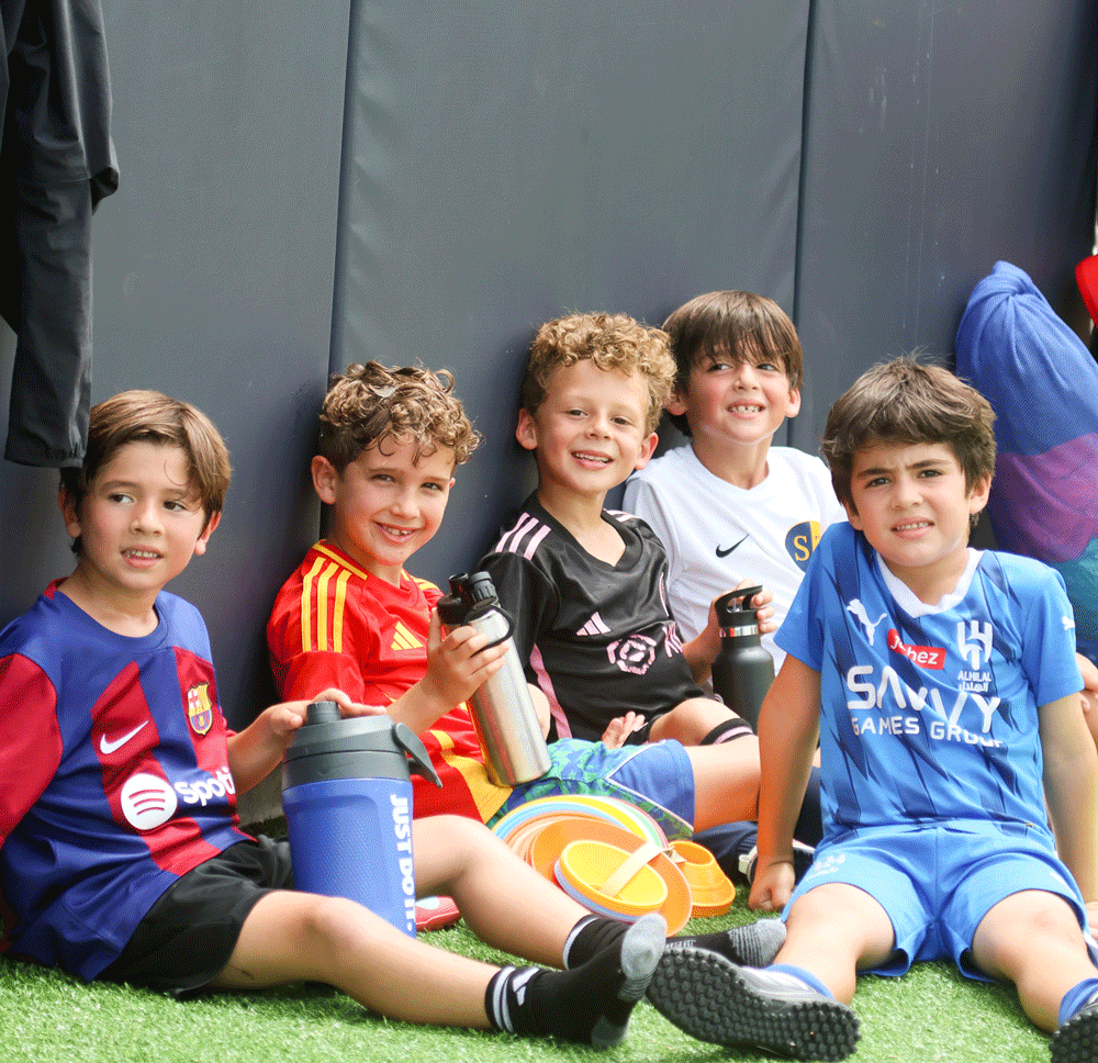 Five young boys in soccer uniforms sit on artificial grass near a wall, smiling at the camera and holding water bottles. Soccer equipment is visible beside them.