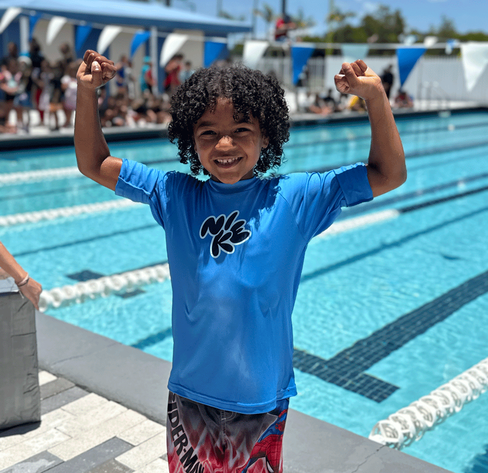 Smiling child in a blue shirt stands by an outdoor swimming pool, flexing both arms, with people and lane dividers visible in the background.