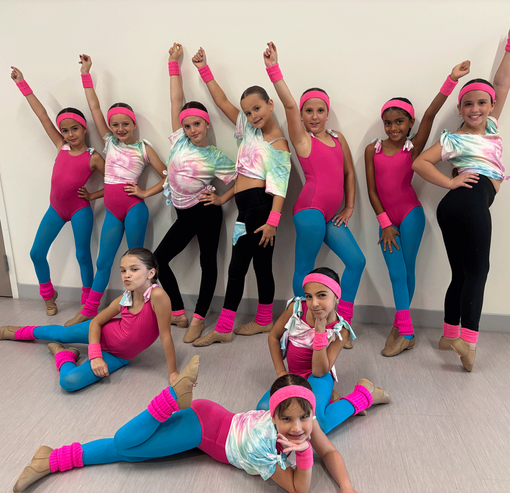 Ten young girls in coordinated dance costumes pose confidently in front of a plain wall, wearing pink, blue, and tie-dye outfits with headbands and matching wrist and ankle bands.