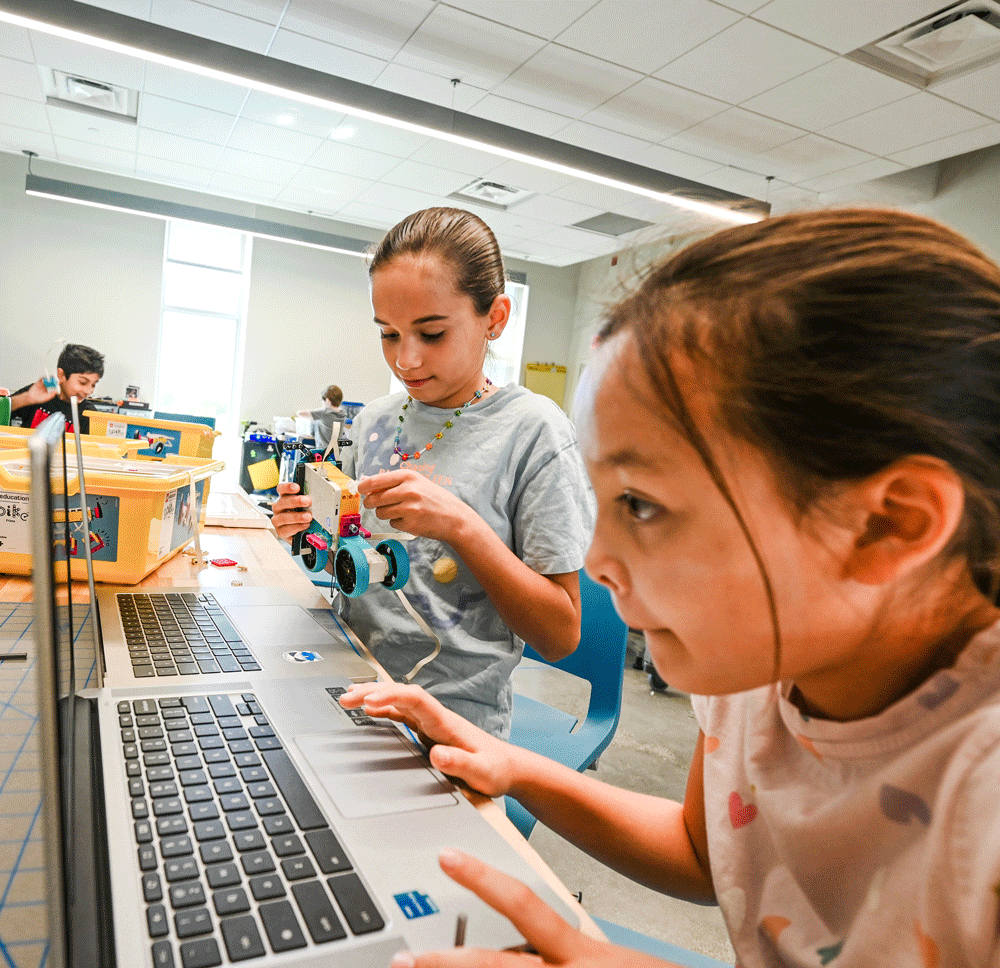 Two children work on a robotics project together at a table, using a laptop and assembling a small robot in a brightly lit classroom.