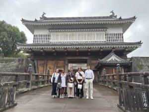 Six people stand on a wooden bridge in front of a traditional Japanese castle gate, posing for a group photo on a cloudy day.
