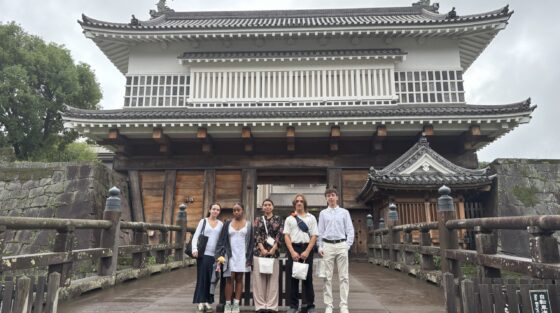 Six people stand on a wooden bridge in front of a traditional Japanese castle gate, posing for a group photo on a cloudy day.