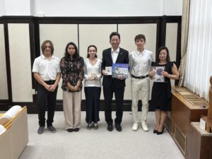 Six people standing indoors, facing the camera. Two hold books, and three others display small objects. The group is dressed in business-casual attire and stands in front of wood-paneled walls.