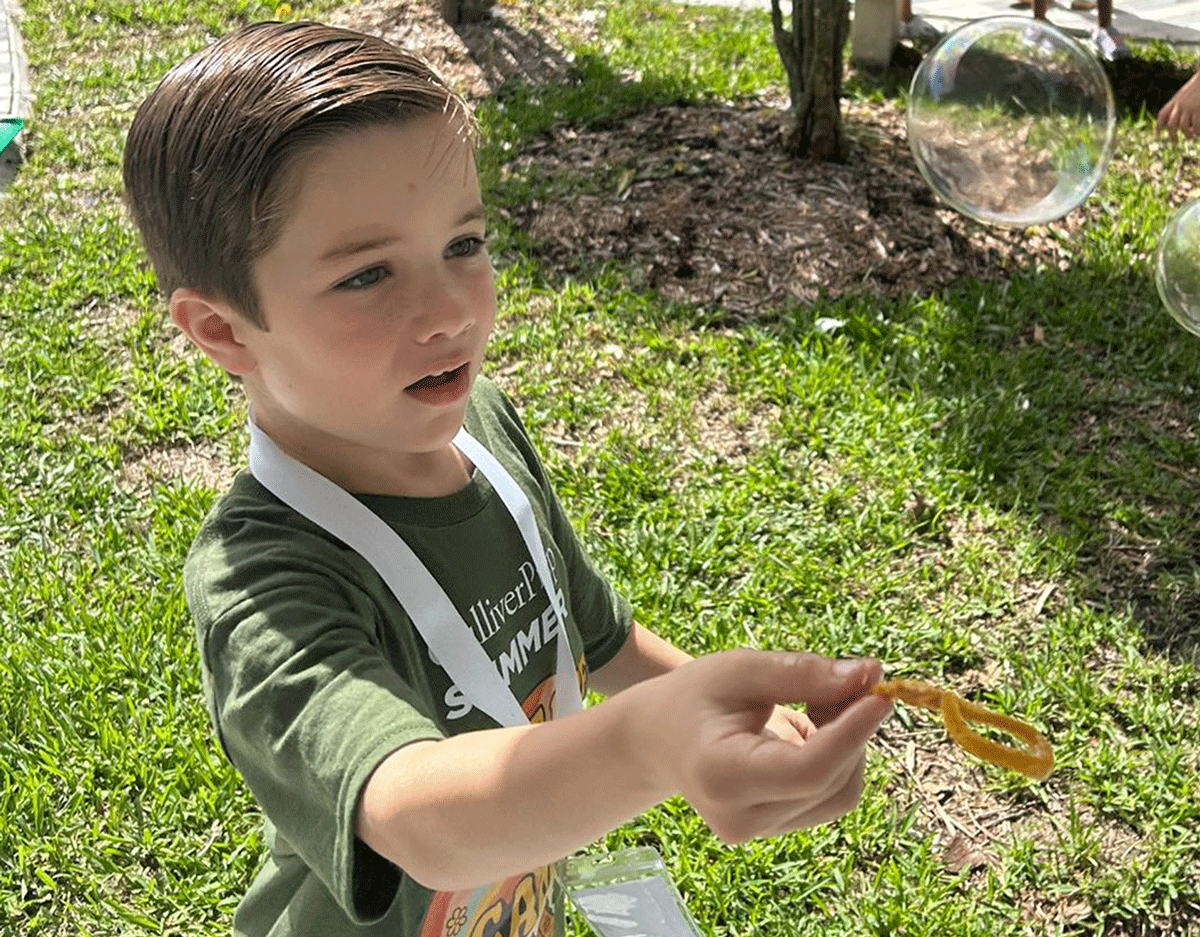 A young boy in a green shirt blows bubbles outdoors on a sunny day, holding a yellow bubble wand with bubbles floating nearby.